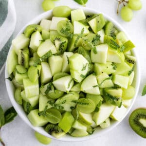 A bowl filled with chopped green fruits including apples, kiwi, grapes, and melon, garnished with fresh mint leaves.