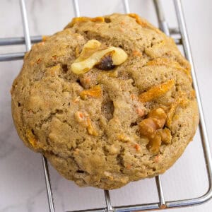 A round, golden-brown cookie with visible carrot shreds and walnut pieces rests on a metal cooling rack.