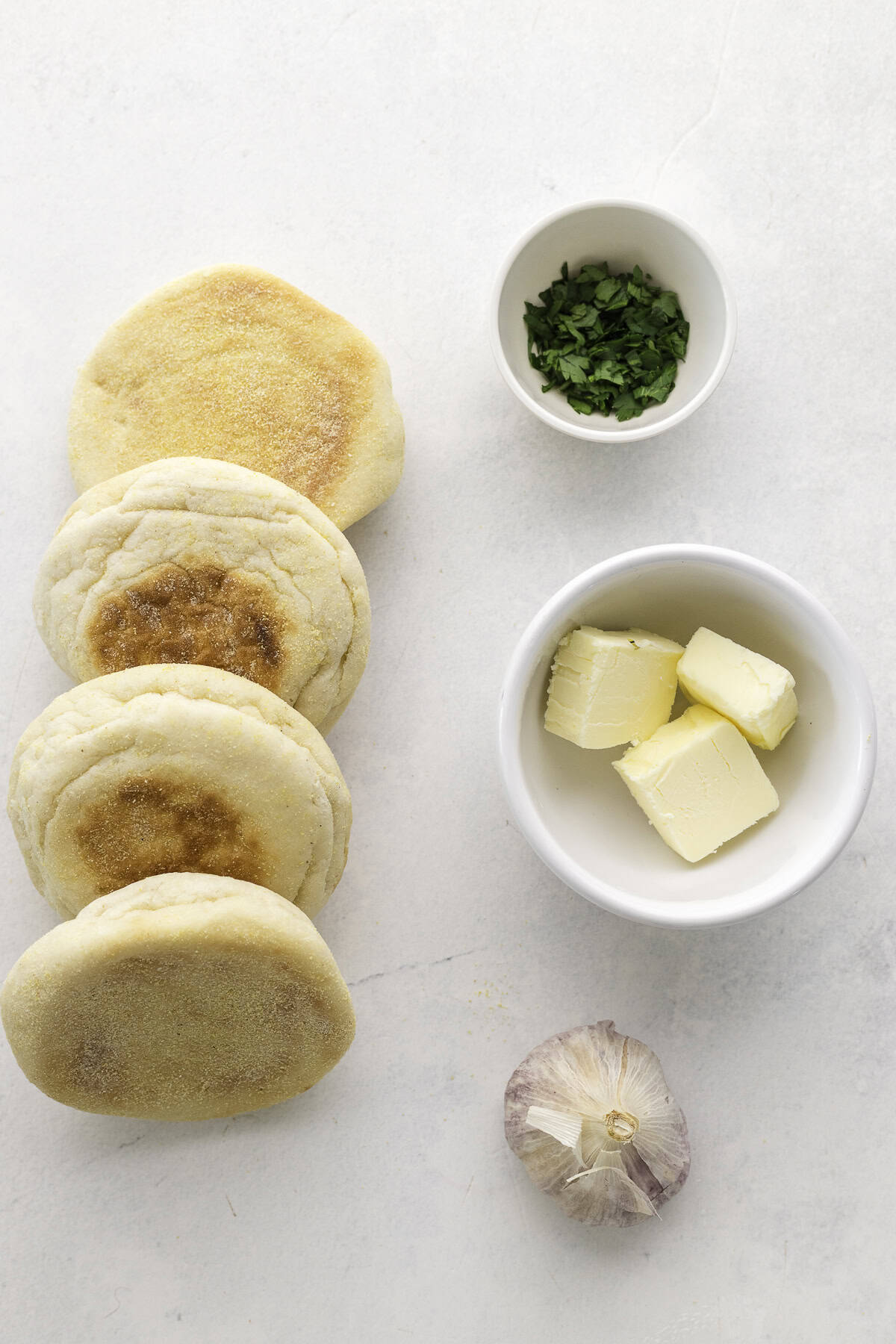 Four English muffins, a small bowl of chopped herbs, a bowl with butter, and a whole garlic bulb arranged on a white surface.
