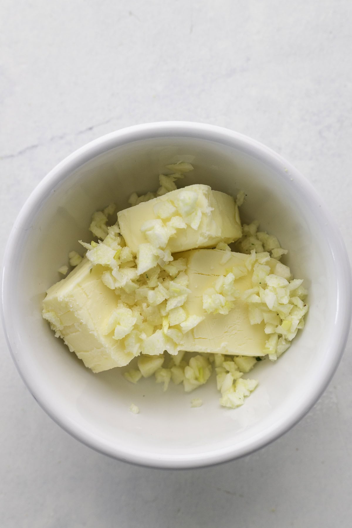 A white bowl containing two slices of butter topped with finely chopped garlic on a light surface.