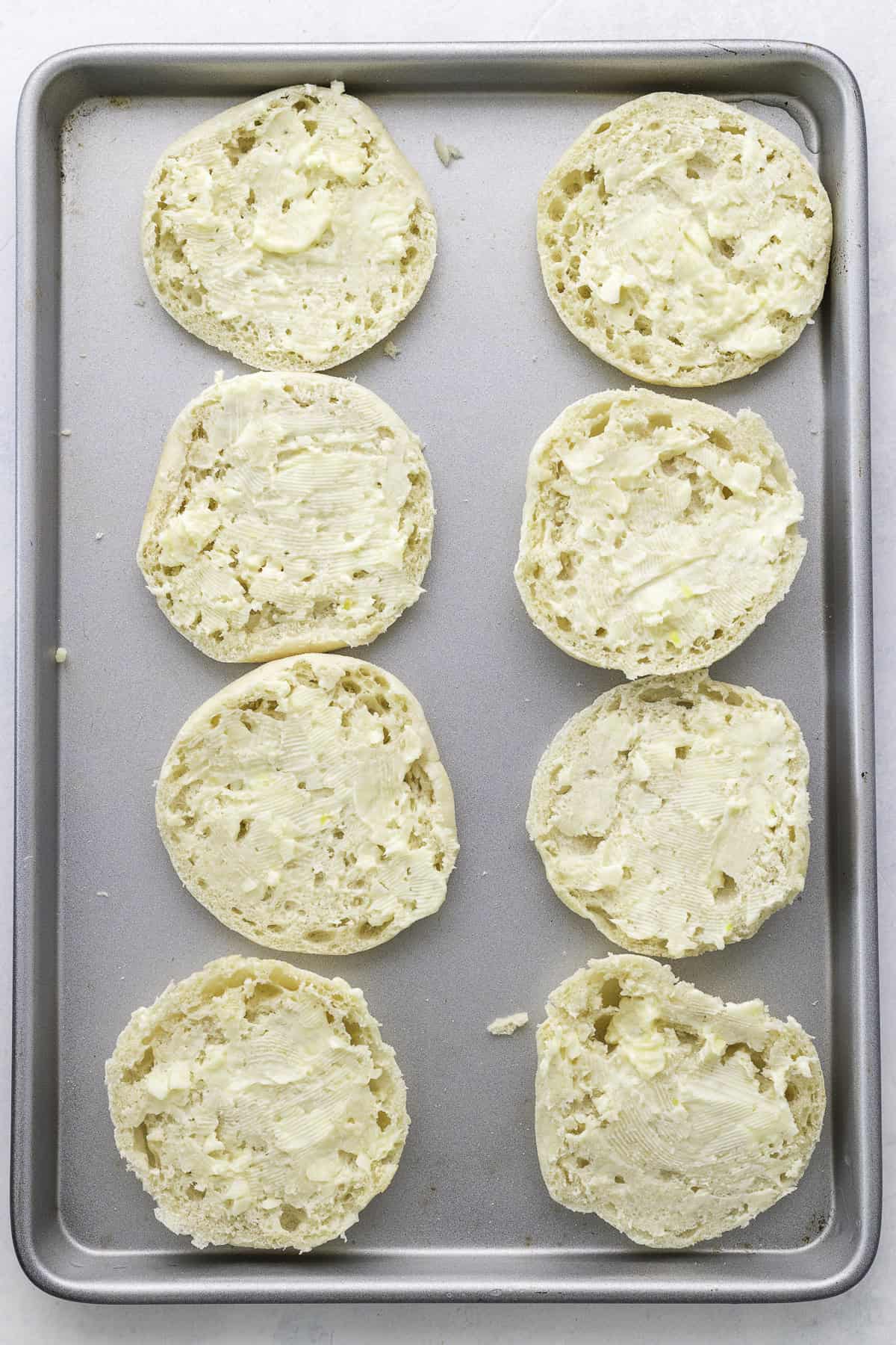 A baking sheet with eight halves of English muffins, each spread with butter and arranged in two rows.