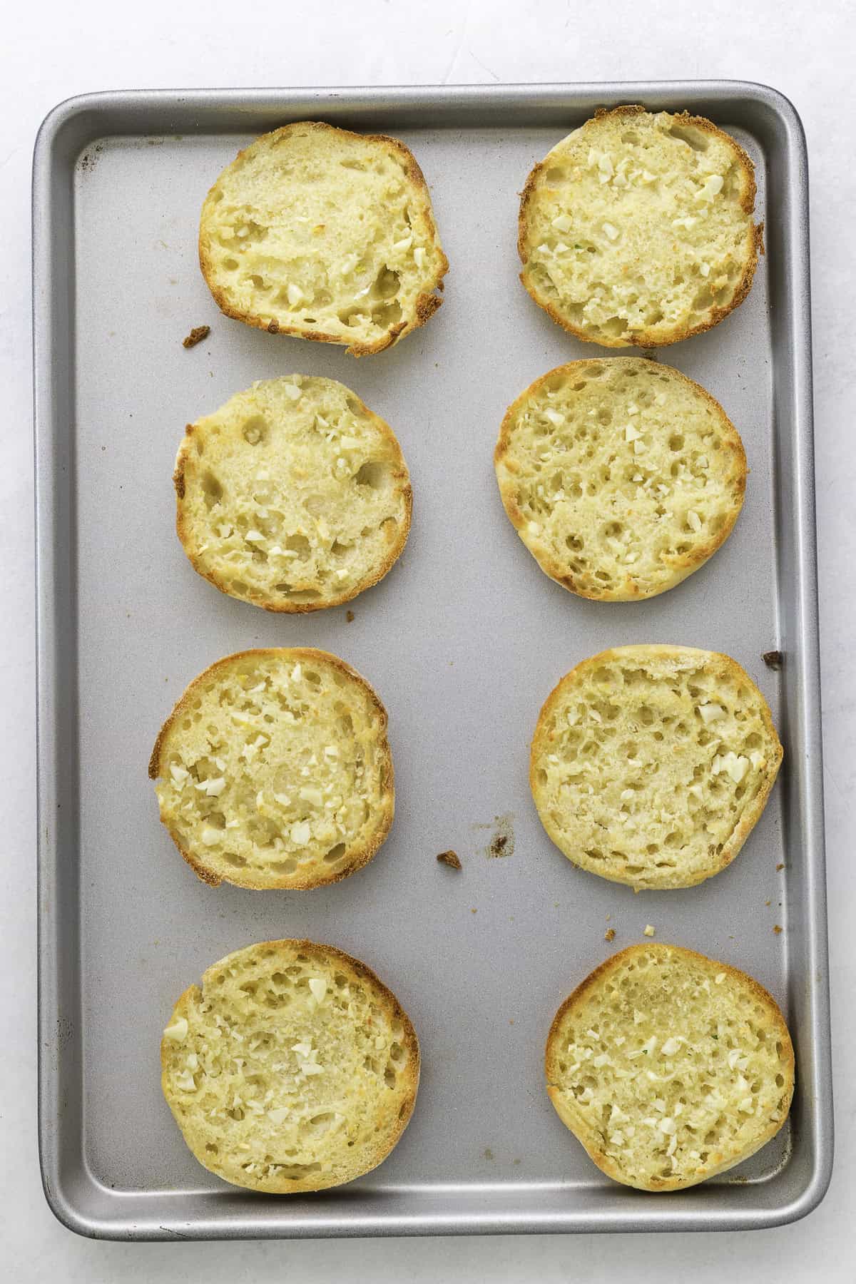 Eight toasted English muffin halves are arranged in two rows on a metal baking sheet.