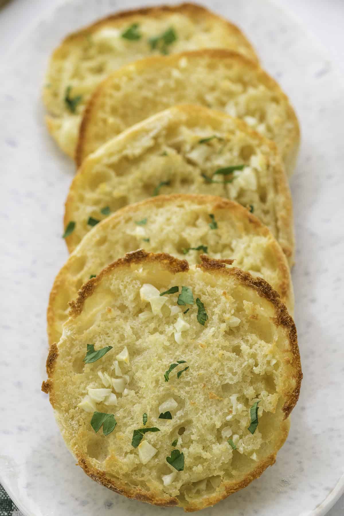 Four slices of toasted english muffin garlic bread topped with chopped garlic and parsley are arranged in a row on a white plate.