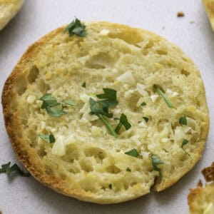 A toasted English muffin garlic bread half topped with minced garlic and chopped parsley, shown on a light-colored surface.