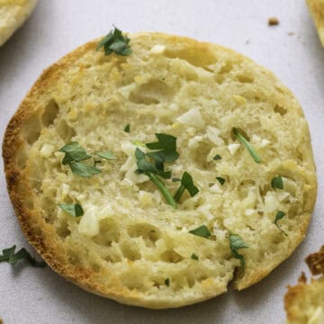 A toasted English muffin garlic bread half topped with minced garlic and chopped parsley, shown on a light-colored surface.