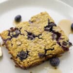 A square piece of baked oatmeal with blueberries on a white plate, drizzled with syrup and garnished with a few whole blueberries.