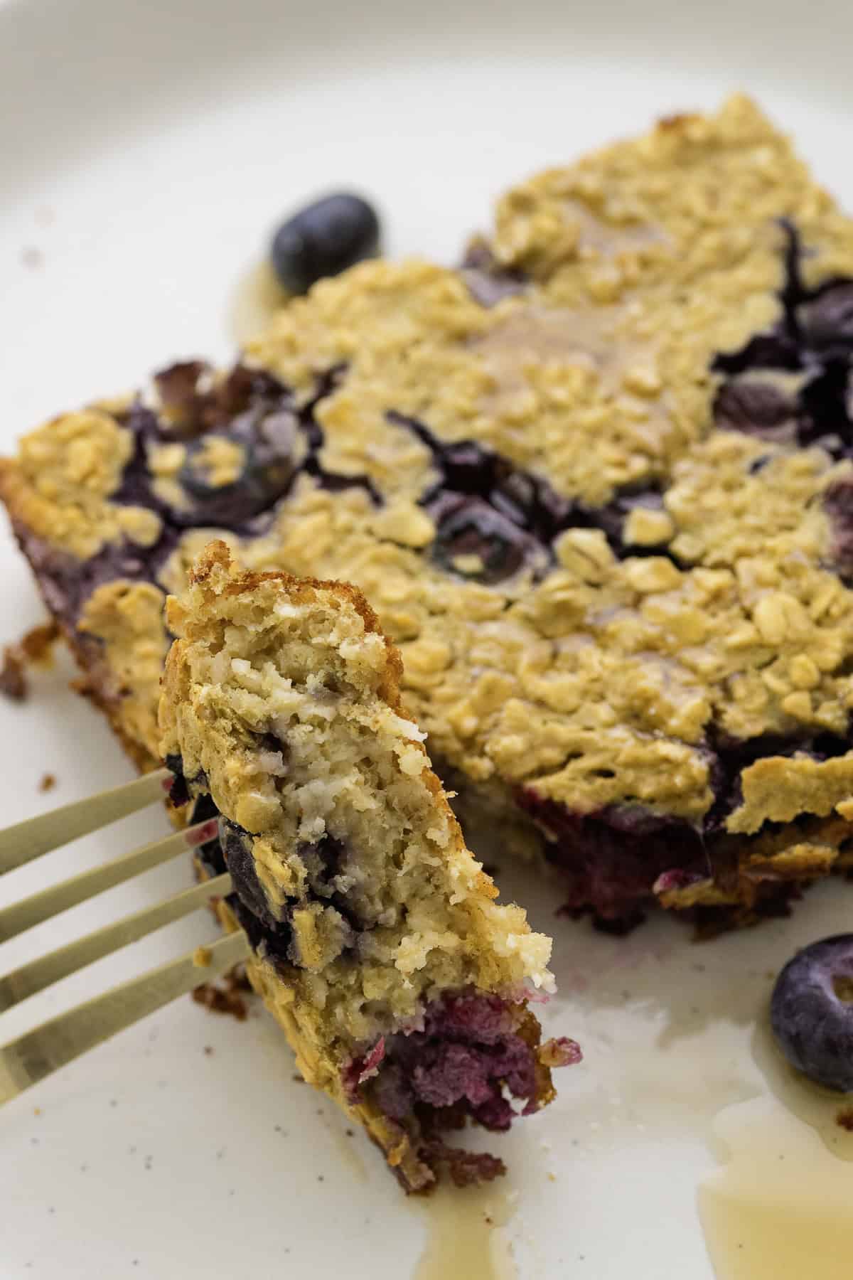 A fork holds a bite of baked oatmeal with blueberries on a plate, with a drizzle of syrup visible in the background.