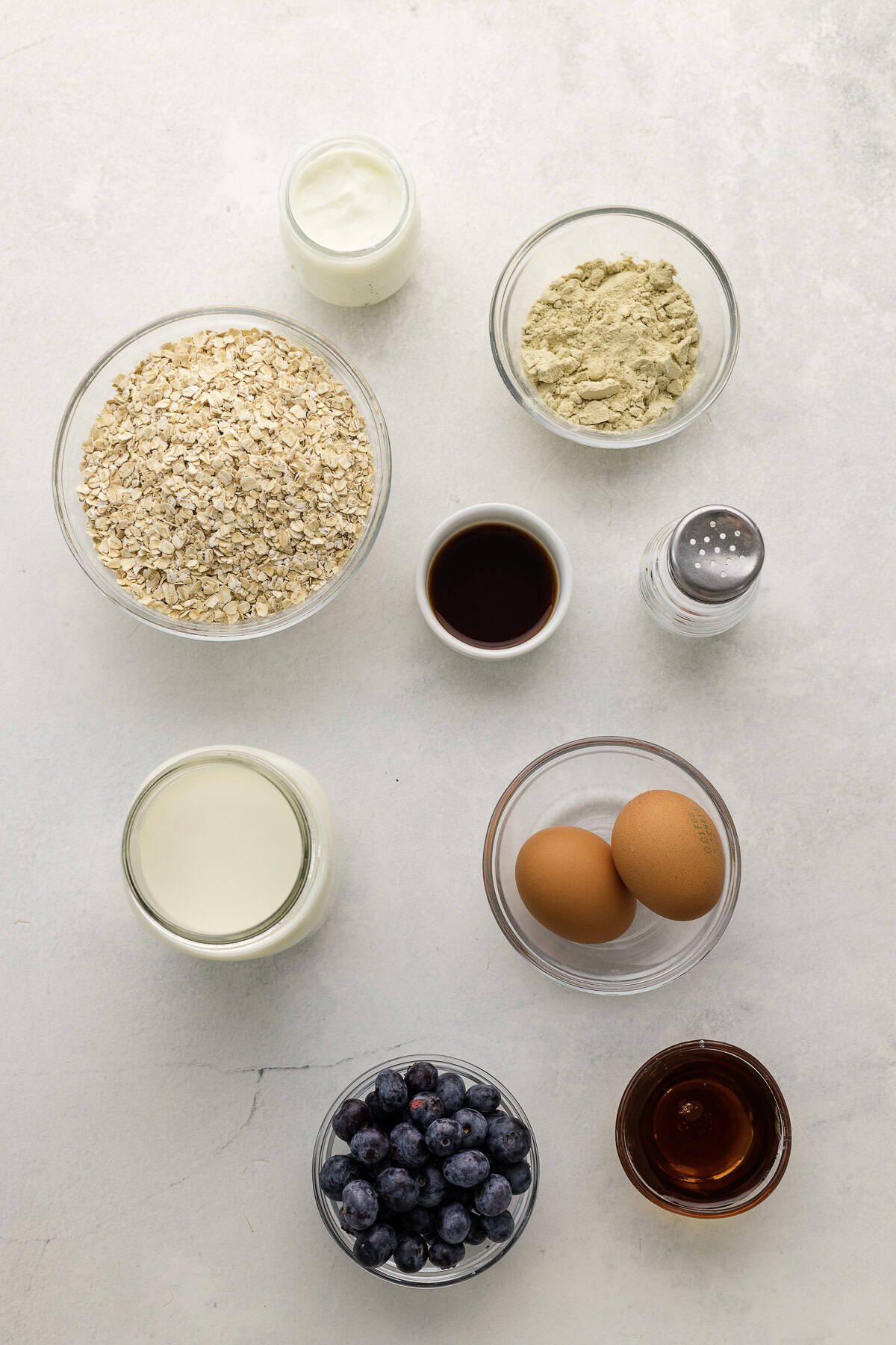 Overhead view of various baking ingredients in bowls and jars, including oats, protein powder, yogurt, milk, eggs, blueberries, vanilla, maple syrup,, and salt.