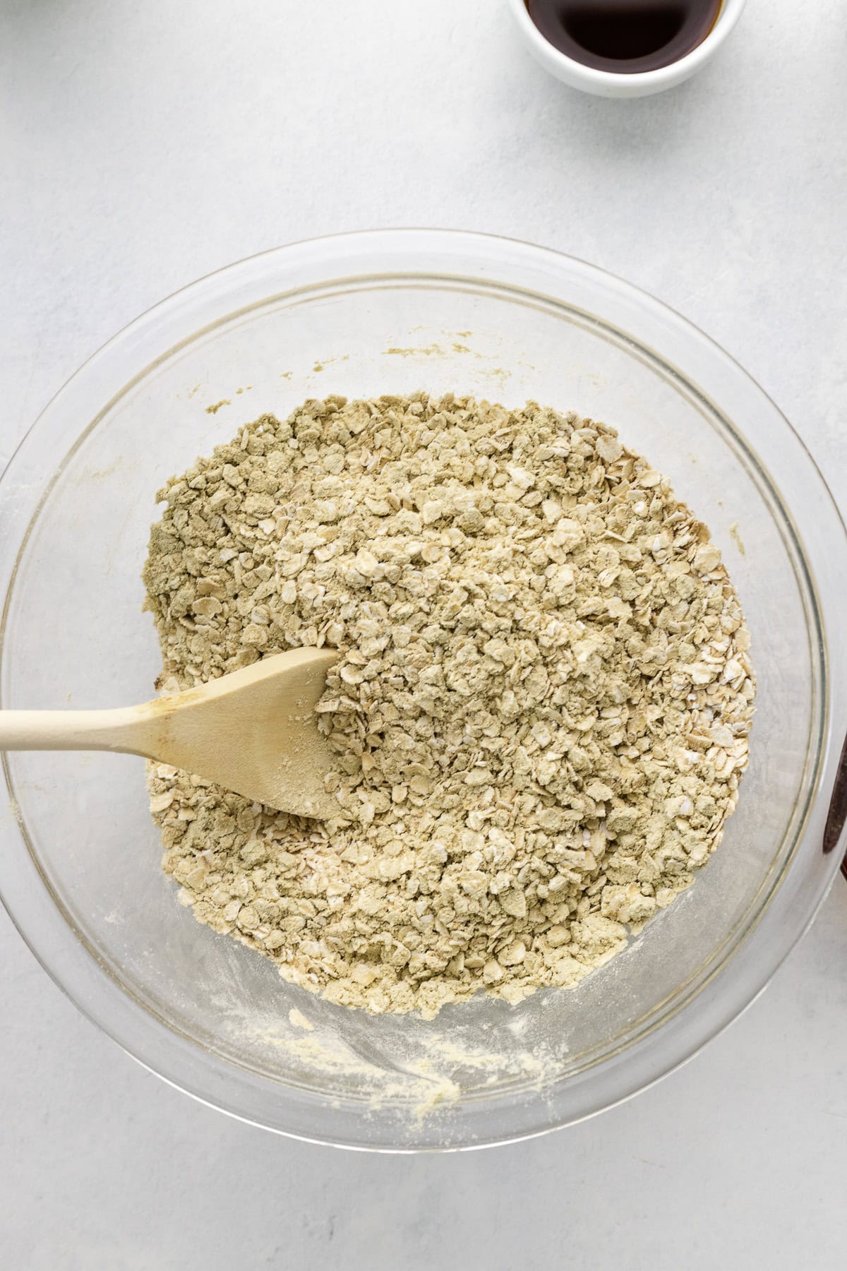 A glass bowl filled with dry oat mixture is being stirred with a wooden spoon; a small cup with dark liquid is partly visible at the top.