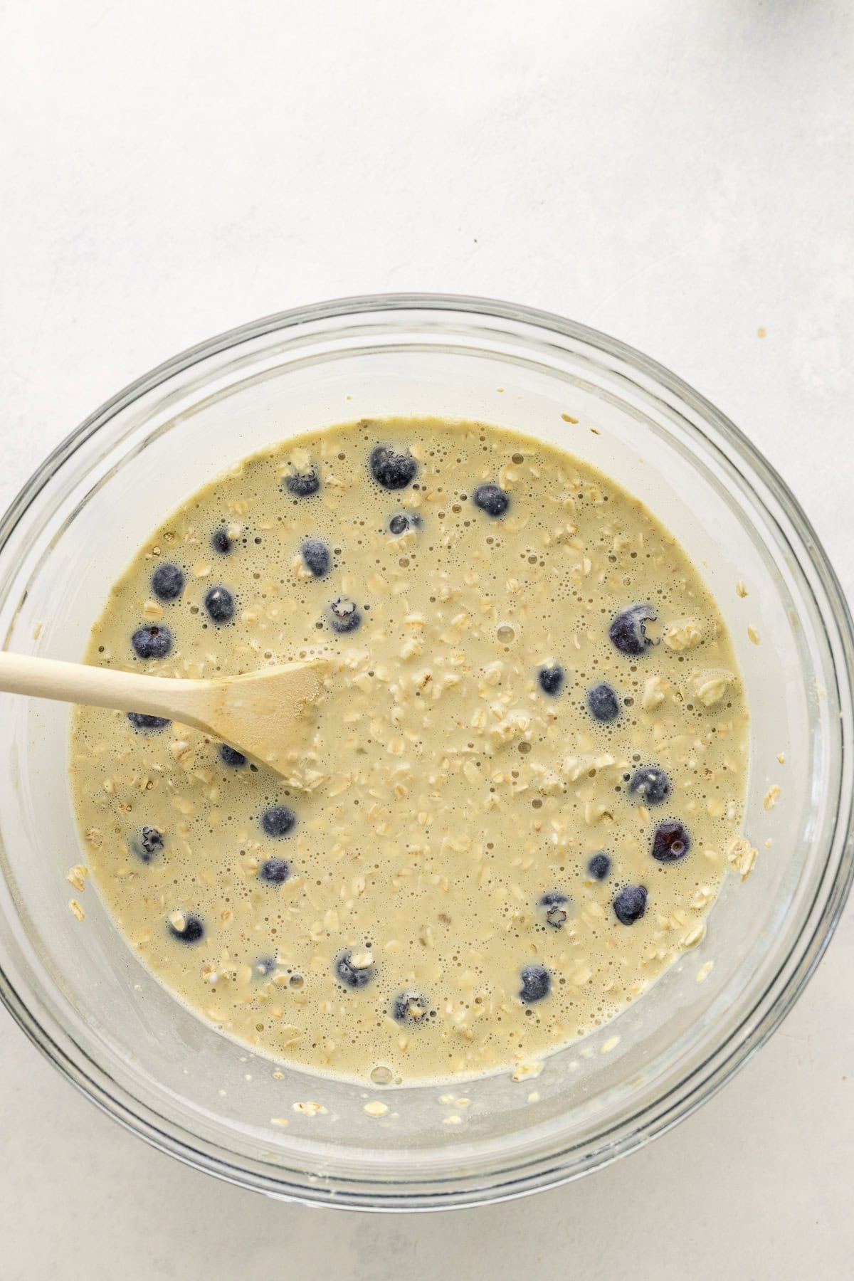 A glass bowl filled with oat and blueberry batter, with a wooden spoon resting inside.