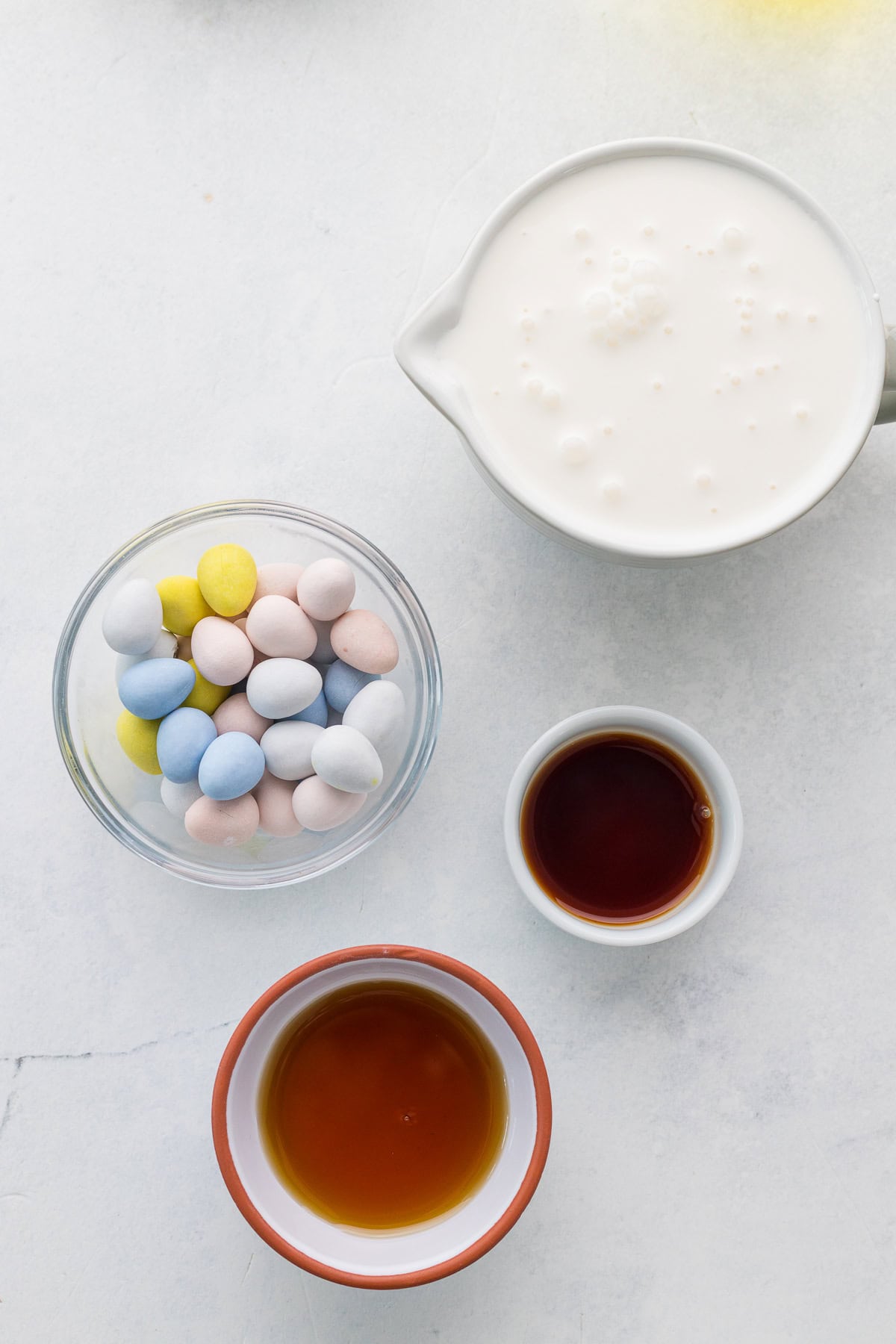 A bowl of candy-coated chocolate eggs, a pitcher of cream, a small bowl of maple syrup, and a small bowl of vanilla set on a white surface.