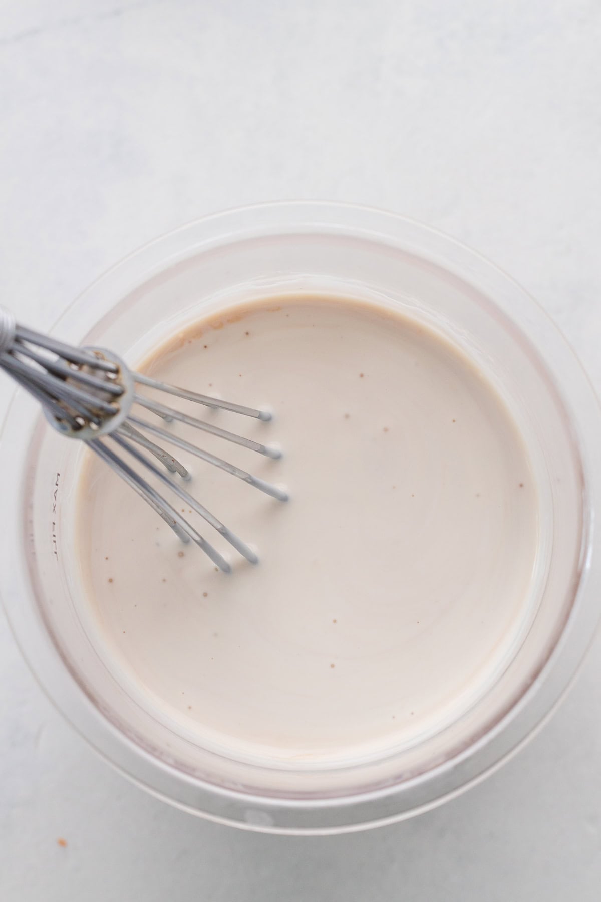 A metal whisk inside a clear bowl of pale, cream mixture, sitting on a light-colored surface.