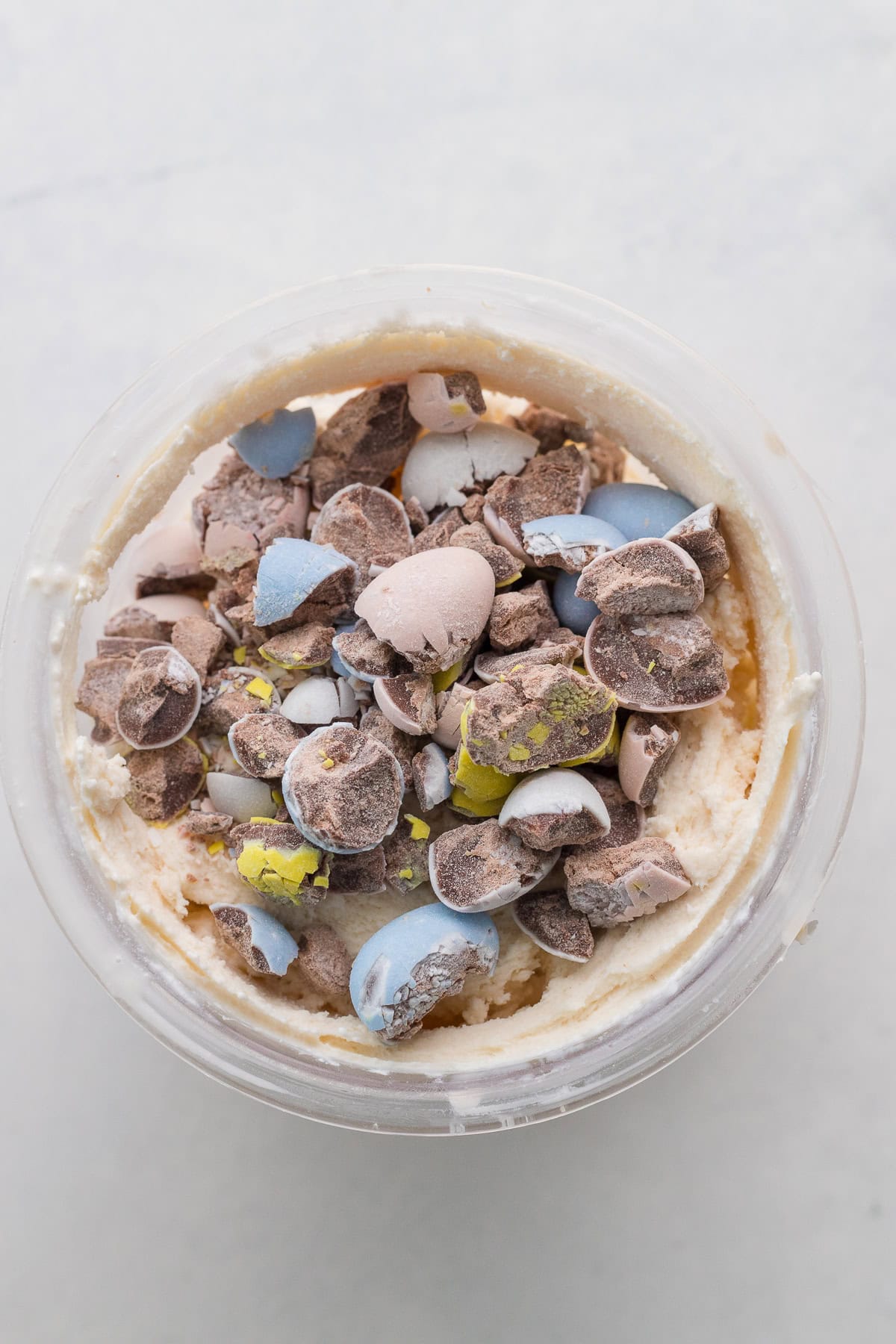 A glass bowl of creamy ice cream topped with crushed candy-coated chocolate pieces in various pastel colors.