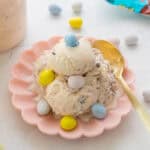 Two scoops of vanilla ice cream with chocolate pieces, topped with colorful candy eggs, served on a pink plate with a gold spoon.