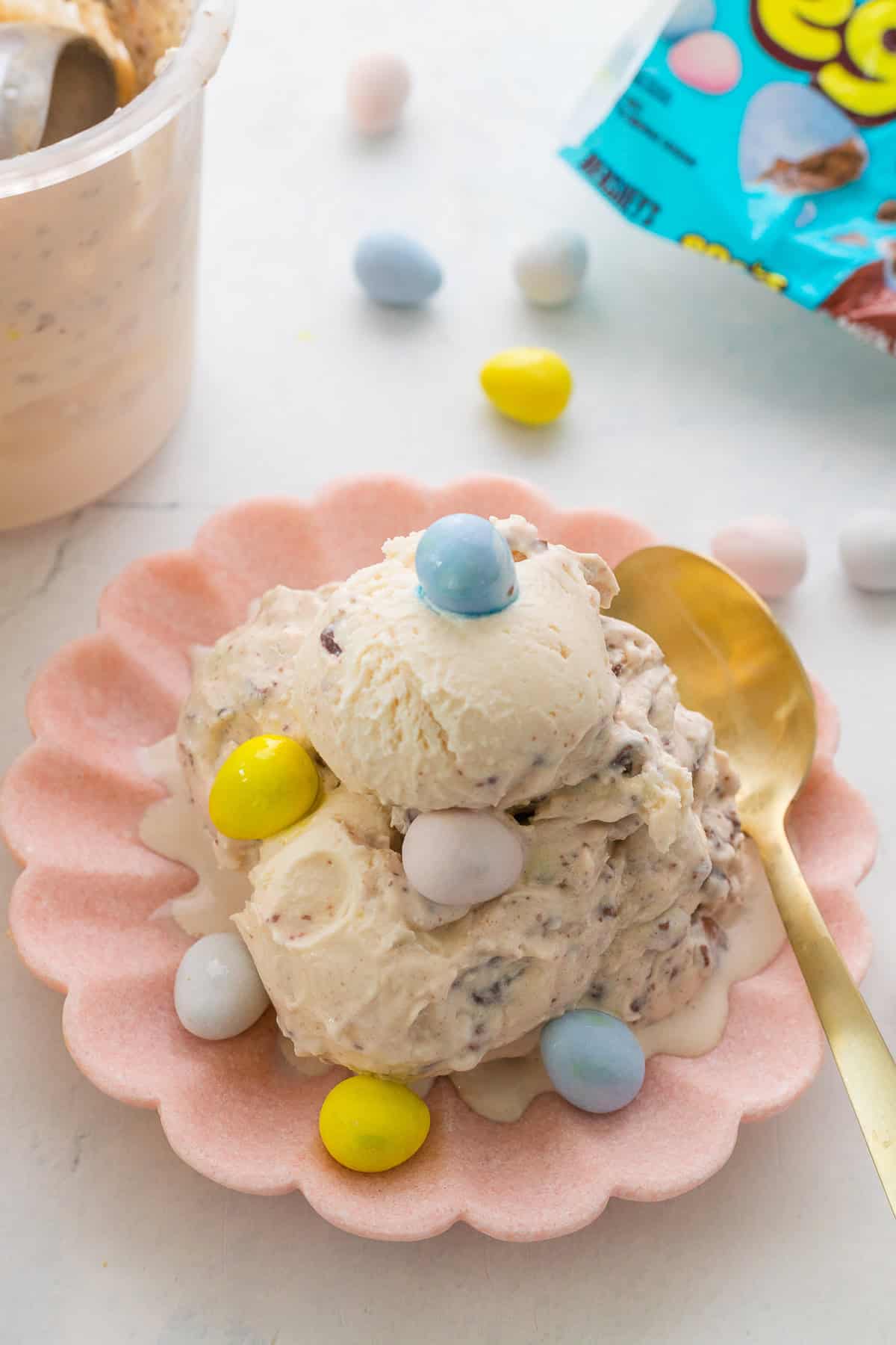 A scoop of mini ggg ice cream with egg chocolate pieces served on a pink plate, topped with pastel candy eggs, next to a gold spoon and a partially opened candy bag.