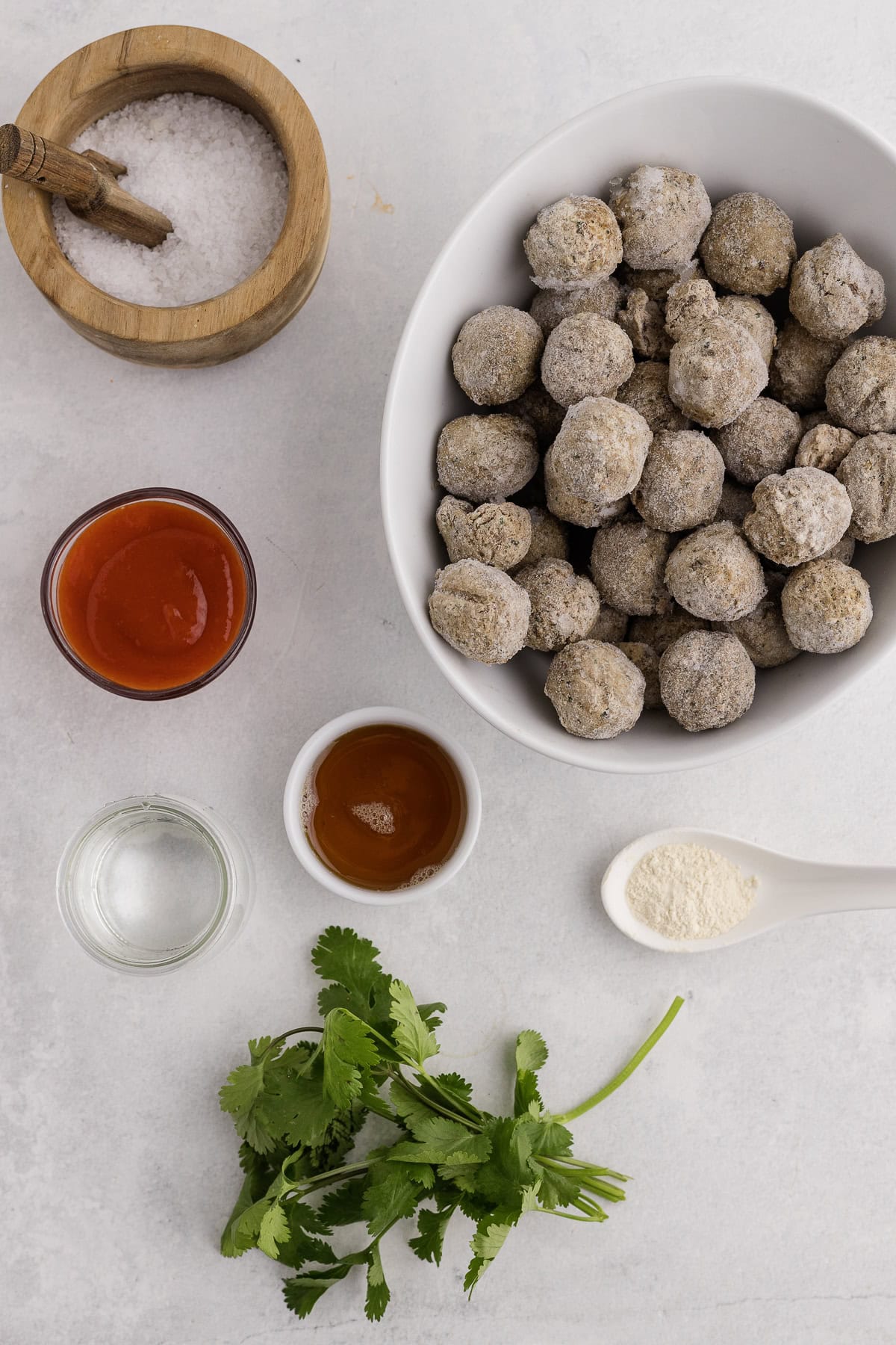 A bowl of frozen meatballs surrounded by small dishes of salt, water, maple syrup, sriracha garlic powder, and fresh cilantro on a light surface.