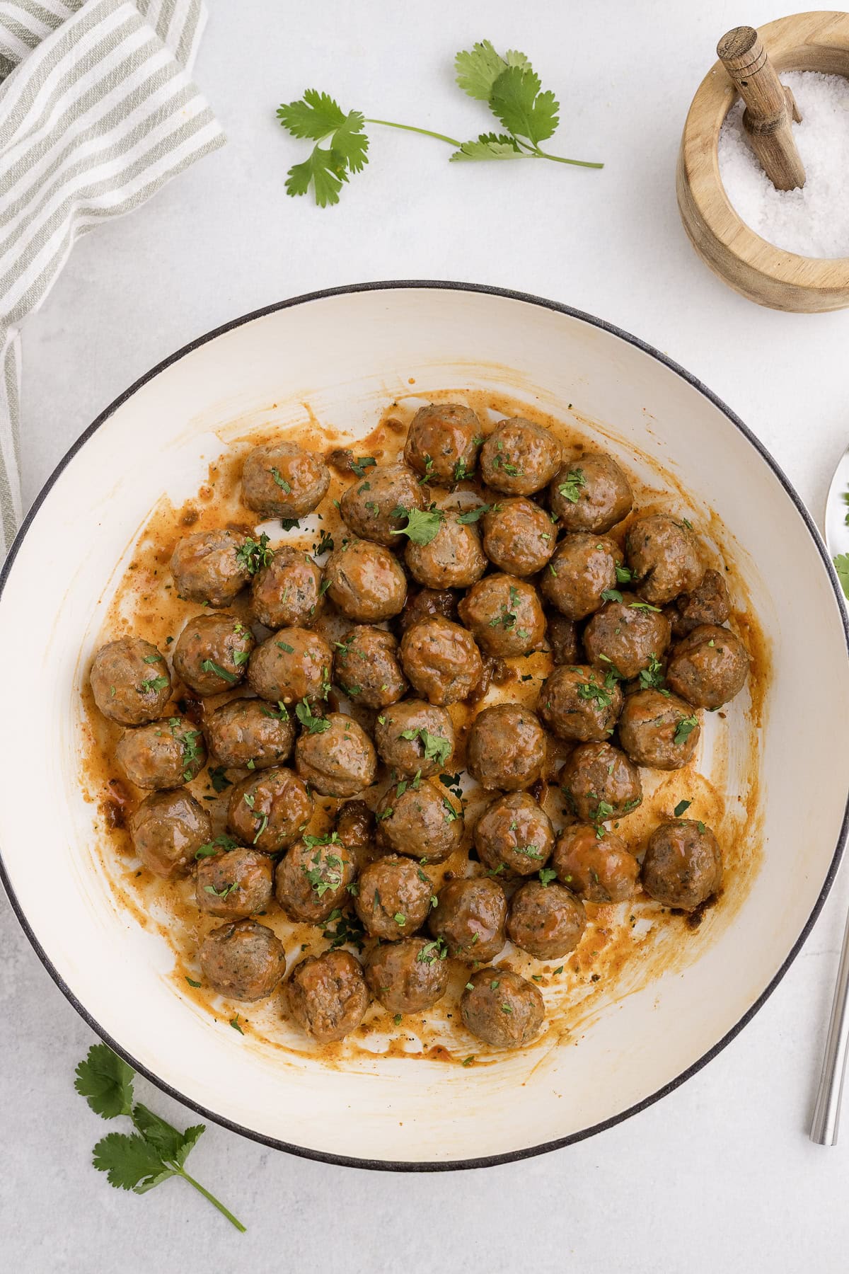 A round white dish filled with sriracha meatballs sauce, garnished with chopped herbs; a striped cloth, fresh cilantro, and a small bowl of salt are nearby.
