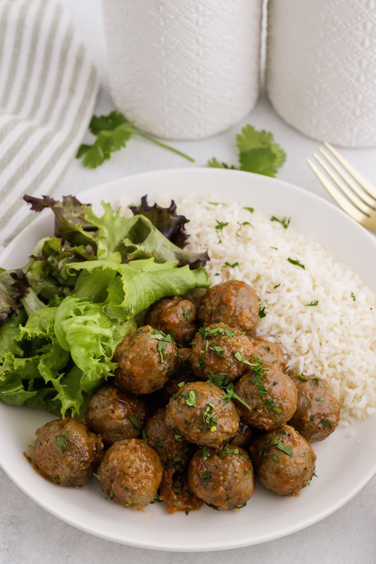 A plate with sriracha meatballs in sauce, white rice, and mixed leafy greens, garnished with chopped herbs.