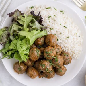 A white plate with a serving of meatballs, steamed white rice, and mixed green salad.