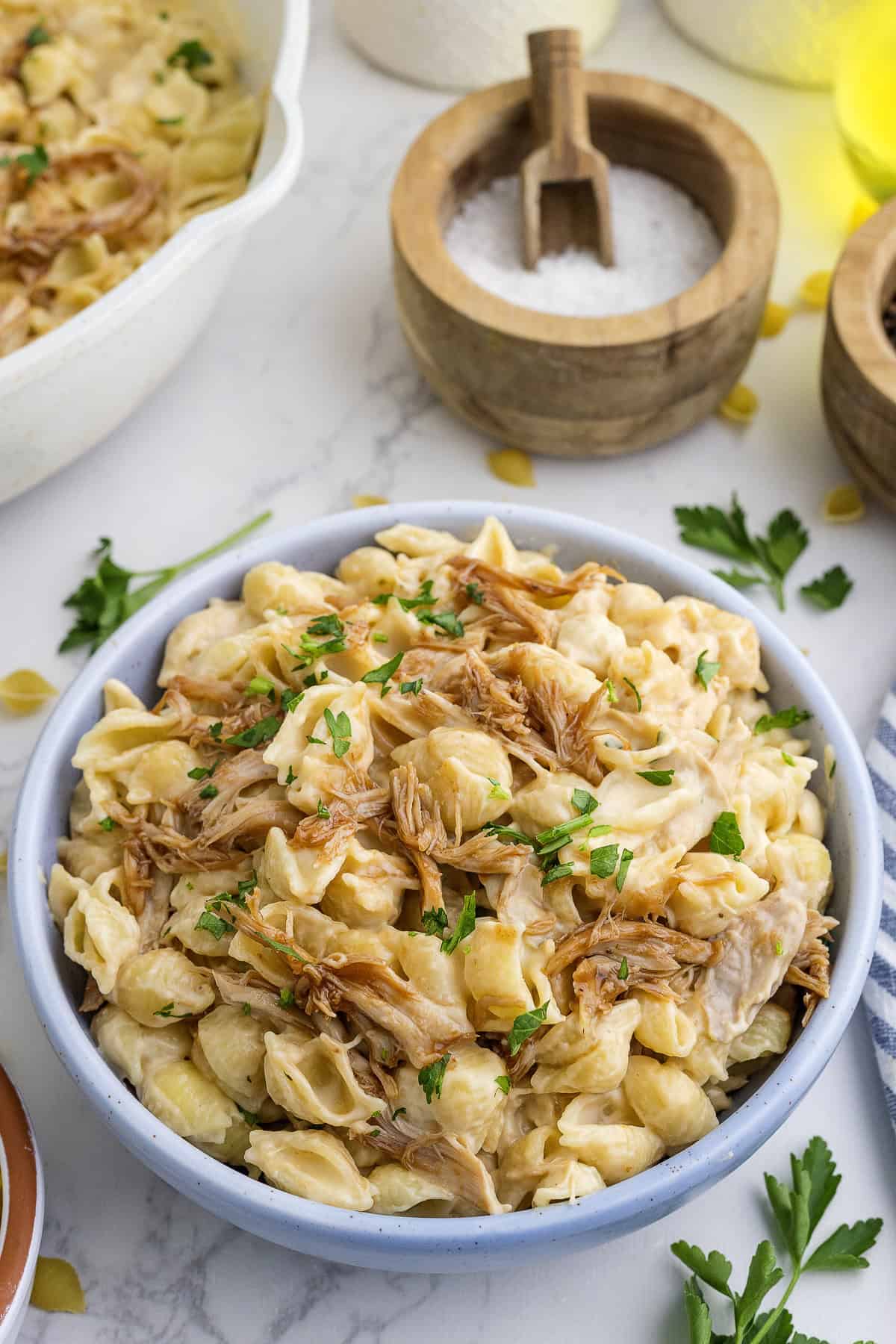 A bowl of creamy pasta shells topped with BBQ shredded chicken and garnished with chopped parsley, placed on a marble surface with salt and parsley nearby.