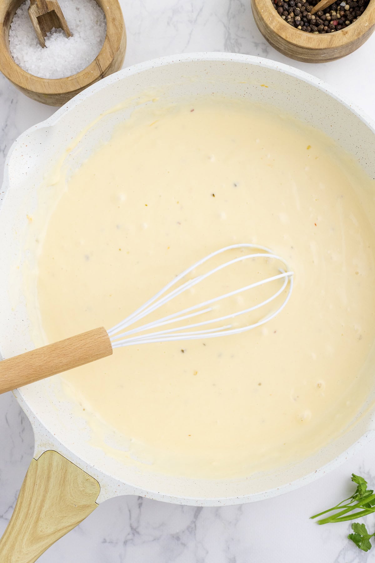 A saucepan filled with creamy cheese sauce, a whisk resting inside. Wooden bowls with salt and pepper sit nearby on a marble surface.