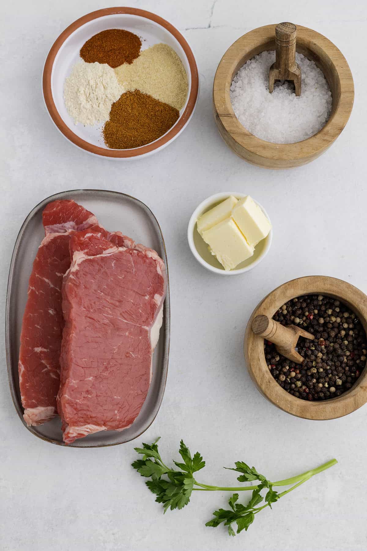 Raw steak on a plate, with bowls of butter, salt, peppercorns, mixed spices, and a sprig of fresh parsley arranged on a white surface.