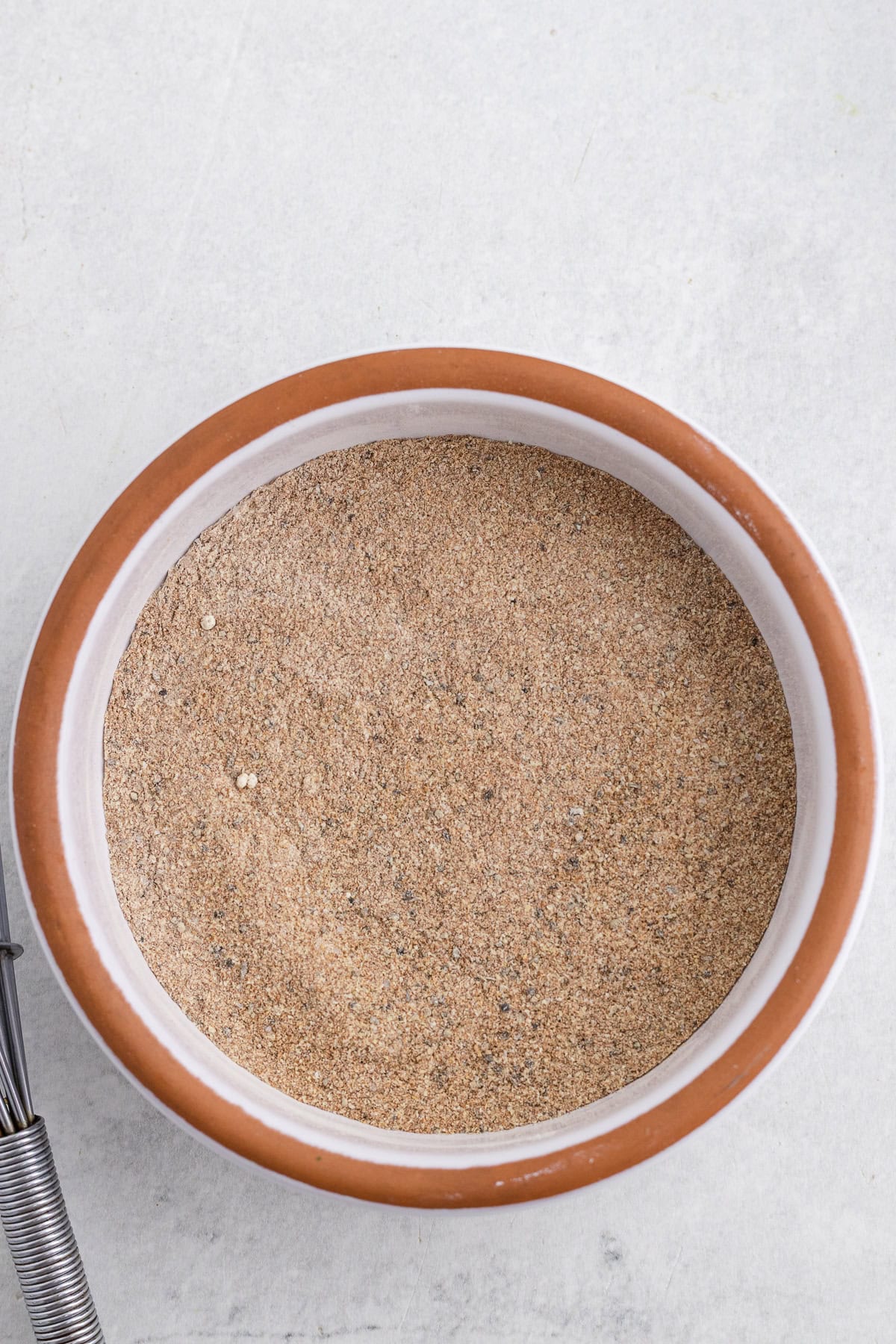 A ceramic bowl filled with a fine, light brown spice mixture sits on a light gray surface, partially visible whisk on the left.