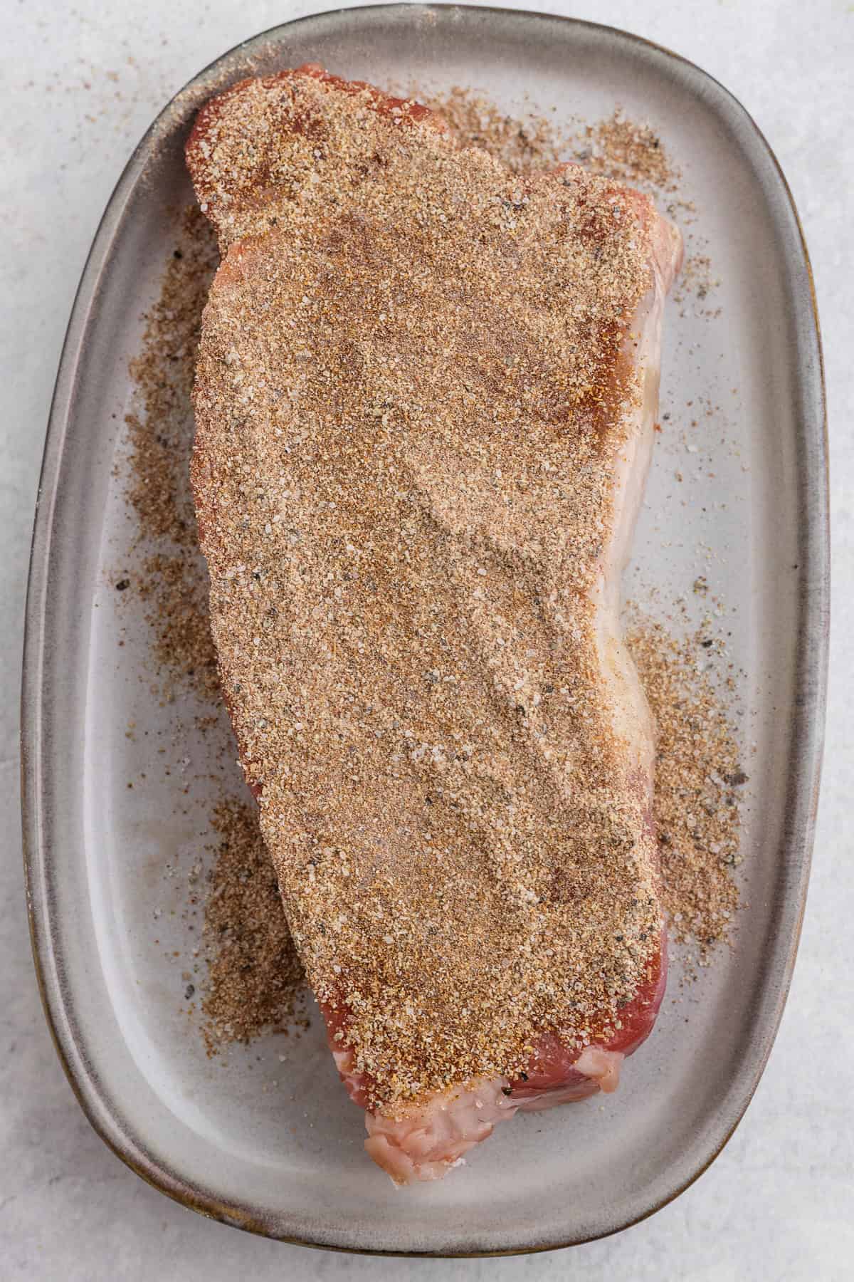 A raw steak on an oval plate is coated with a layer of dry seasoning rub.