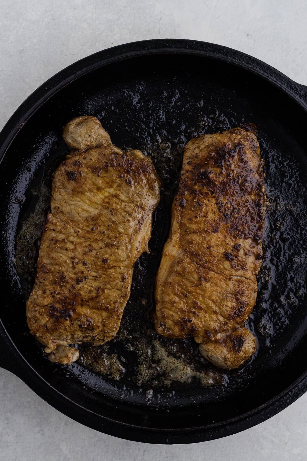 Two seared steaks cooking in a black cast iron skillet, with browned surfaces and some melted fat visible.