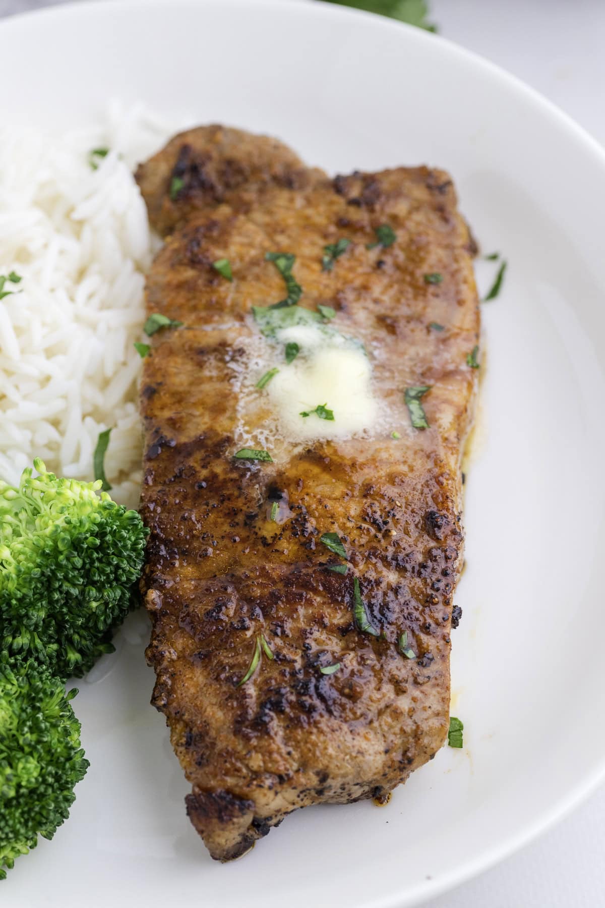 A cooked blackened steak topped with melted butter and herbs, served with white rice and steamed broccoli on a white plate.