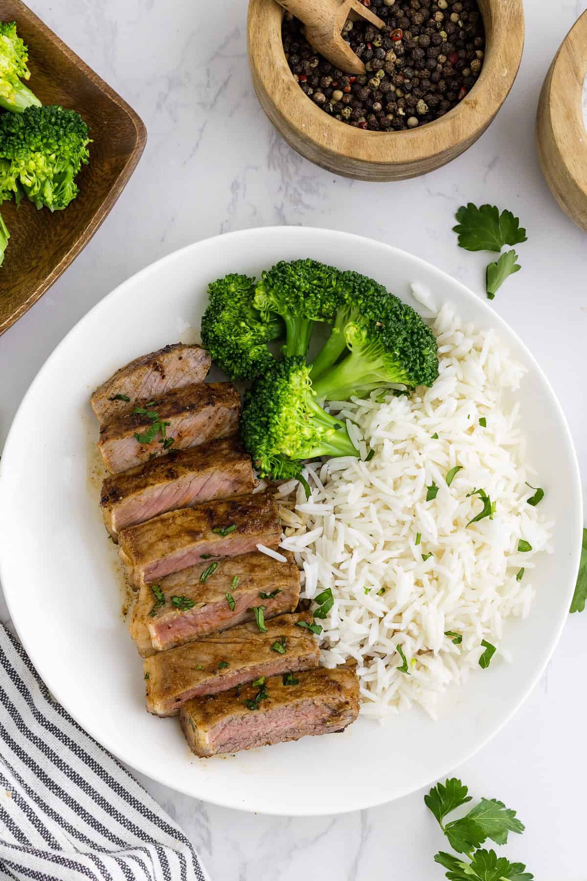 A white plate with sliced blackened steak, steamed broccoli, and white rice, garnished with parsley, on a marble surface.