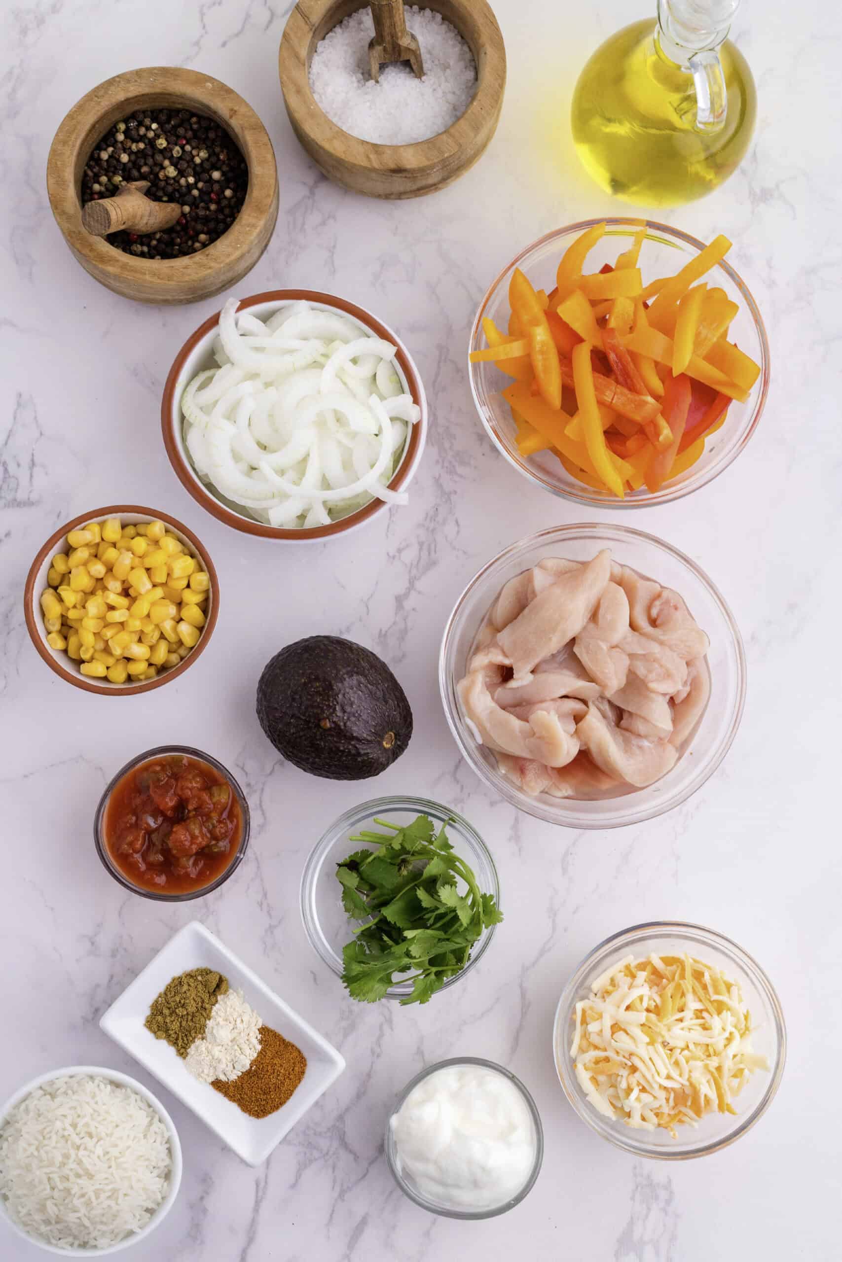 Vue de haut en bas d'un assortiment d'ingrédients sur une surface en marbre, comprenant des tranches de poivrons, d'oignons, de maïs, d'avocat, de poulet, de salsa, d'herbes, d'épices, de fromage, de riz, de crème sure, d'huile, de sel et de poivre.