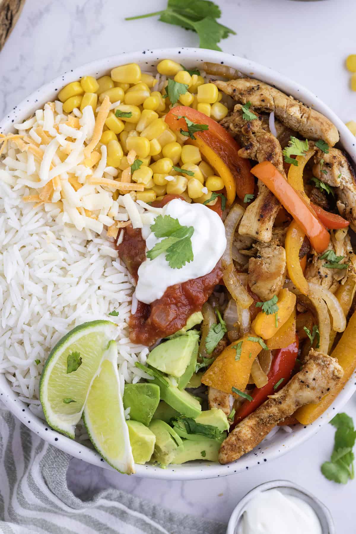 Un bol garni de riz blanc, de maïs, de fromage râpé, de poulet grillé aux poivrons et oignons, d'avocat, de quartiers de citron vert, de salsa et de crème fraîche, le tout parsemé de coriandre.