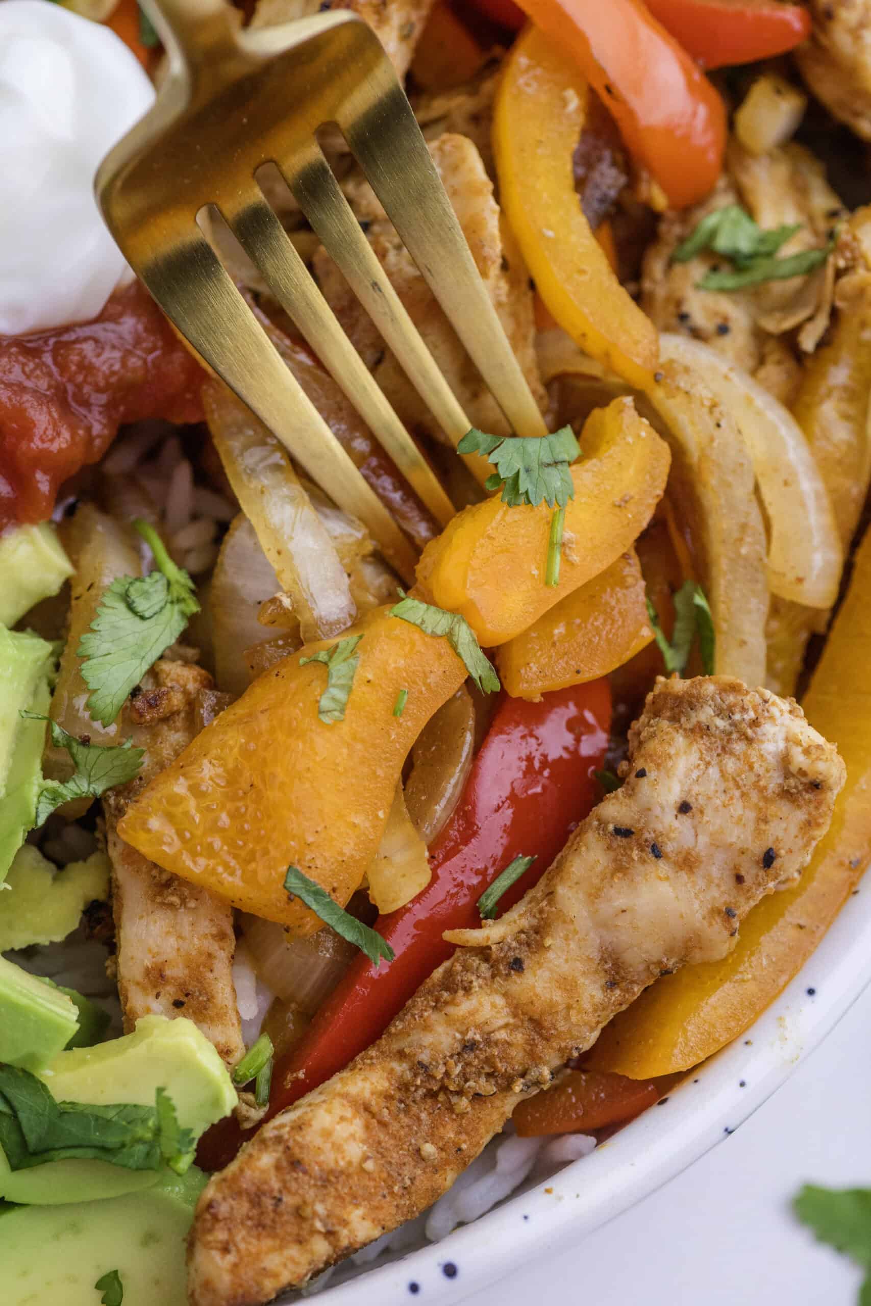 Gros plan d'une fourchette dans un bol rempli de lamelles de poulet assaisonnées, de tranches de poivrons, d'oignons, d'avocats, de salsa et de coriandre fraîche.