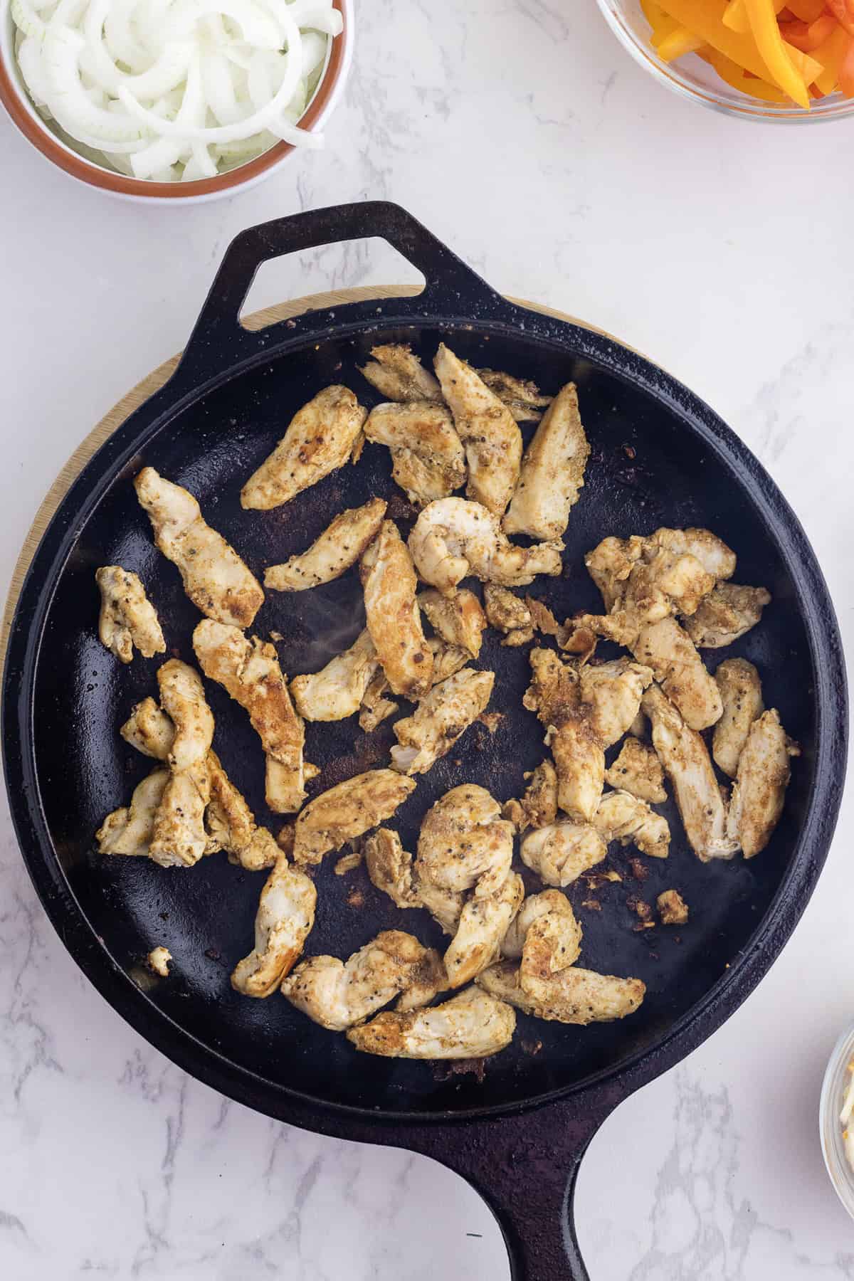 Des lamelles de poulet assaisonnées cuisent dans une poêle en fonte noire, avec des bols d'oignons et de poivrons émincés sur une surface blanche.