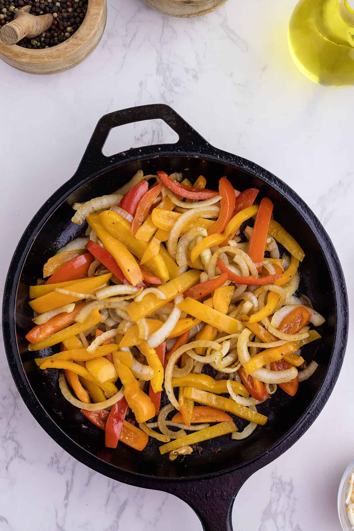Sliced yellow and red bell peppers with onions are sautéing in a black cast iron skillet on a white surface.