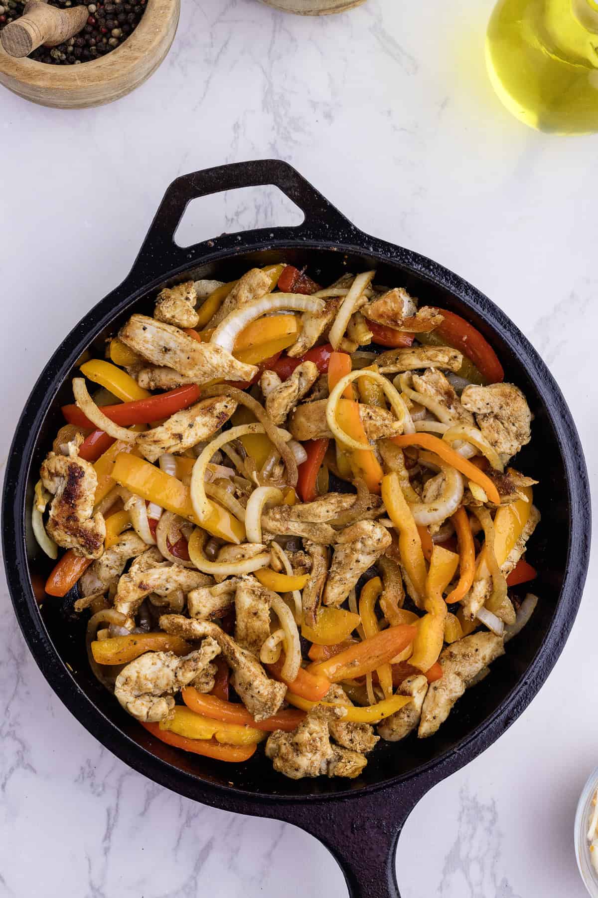 Une poêle en fonte remplie de lanières de poulet cuites, de tranches de poivrons jaunes et rouges et d'oignons sur une surface en marbre blanc.