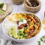 A bowl of rice topped with grilled chicken, sautéed peppers, corn, avocado, salsa, sour cream, shredded cheese, and lime wedges, garnished with cilantro.