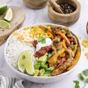 Un bol rempli de riz, de poulet grillé, de poivrons sautés, de maïs, d'avocat, de salsa, de fromage râpé, de quartiers de citron vert et d'une cuillerée de crème aigre, le tout garni de coriandre.