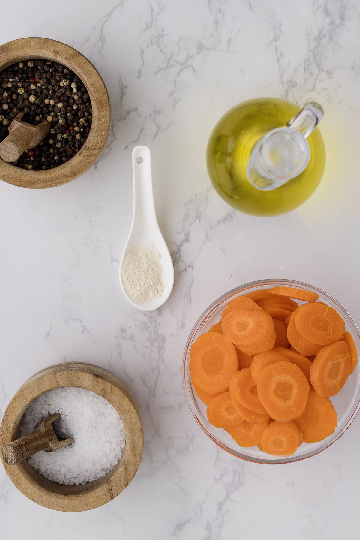 Un bol de carottes coupées en rondelles, une carafe d'huile d'olive, un bol de poivre en grains, un bol de sel et une cuillère d'ail en poudre sont disposés sur une surface en marbre blanc.