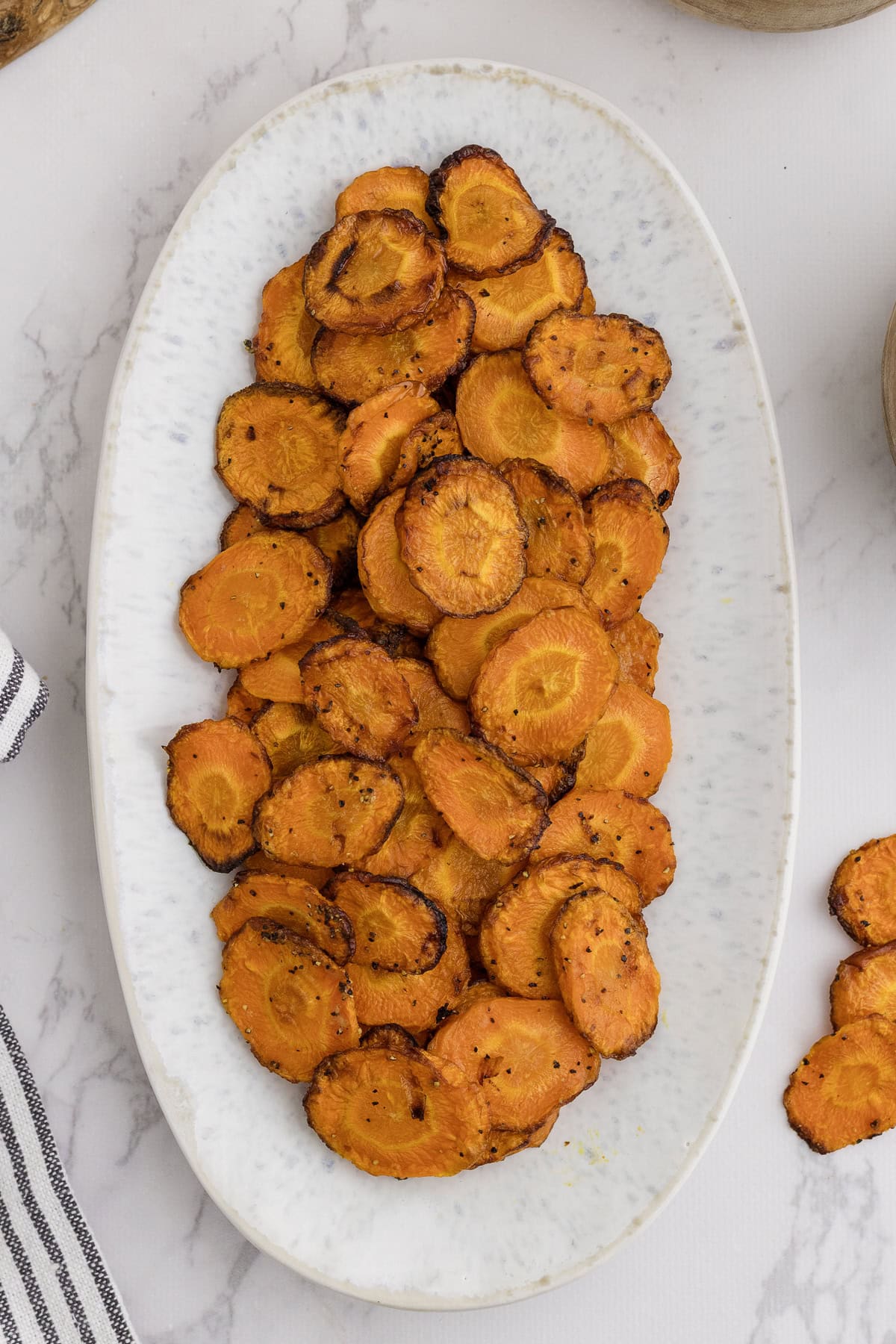Une assiette ovale blanche contient une pile de chips de carottes grillées avec assaisonnement, sur une surface en marbre.