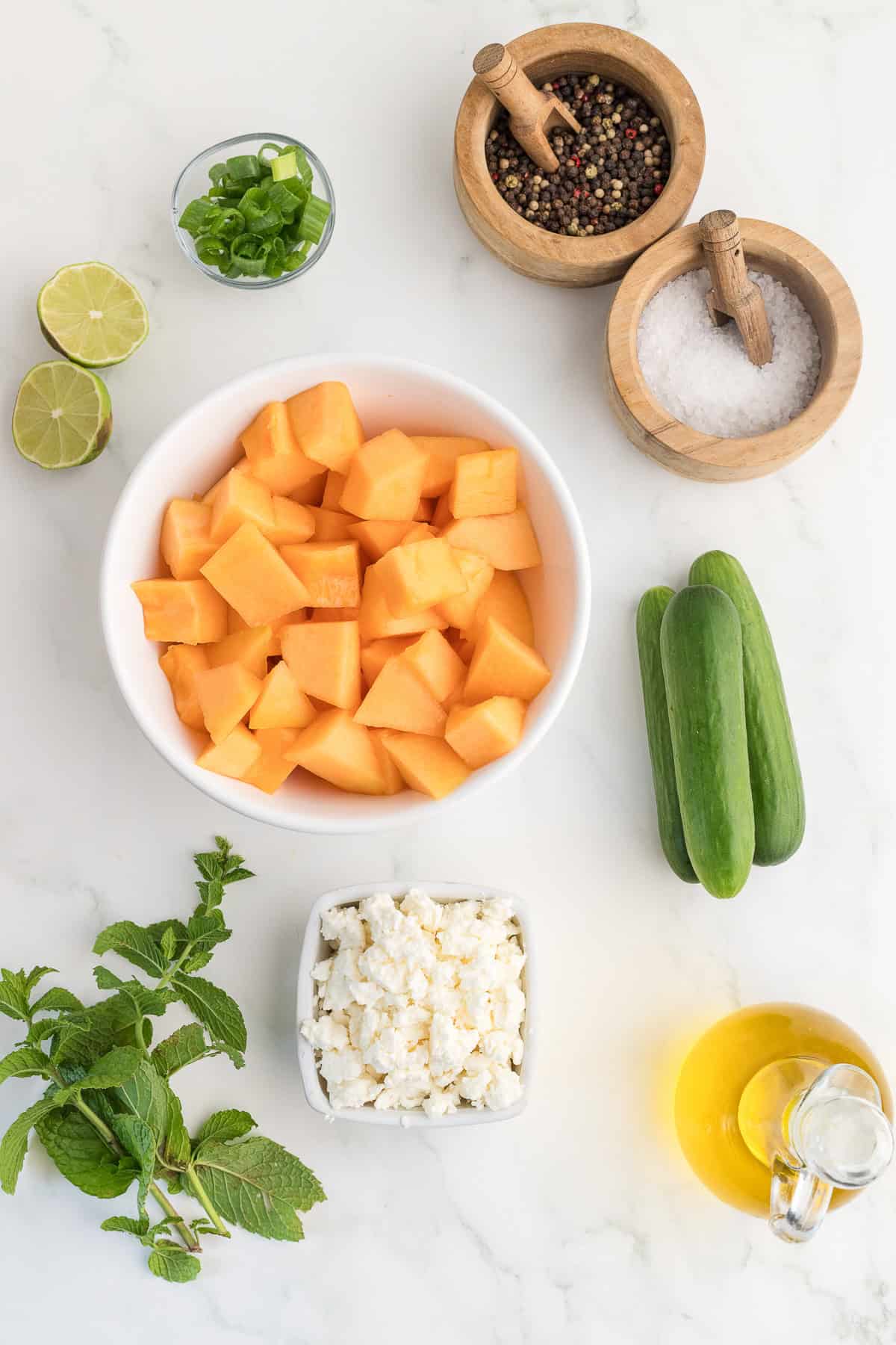 A bowl of cubed cantaloupe, cucumbers, feta cheese, fresh mint, lime halves, green onions, olive oil, salt, and pepper on a white surface.