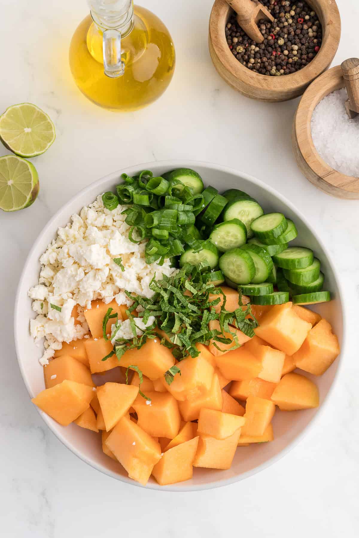 A bowl with cubed cantaloupe, chopped cucumber, feta cheese, green onion, and mint, next to olive oil, halved lime, peppercorns, and a bowl of salt on a white surface.