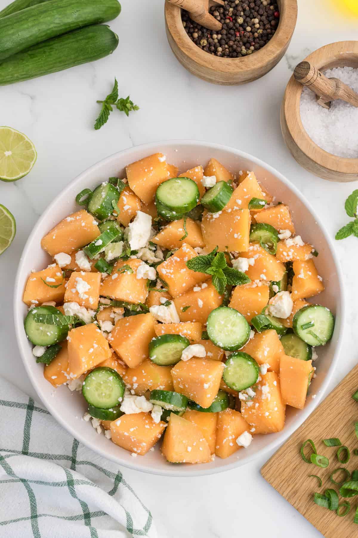A bowl of salad with cantaloupe chunks, cucumber slices, crumbled feta cheese, and mint leaves on a white surface with lime, a towel, salt, and pepper nearby.