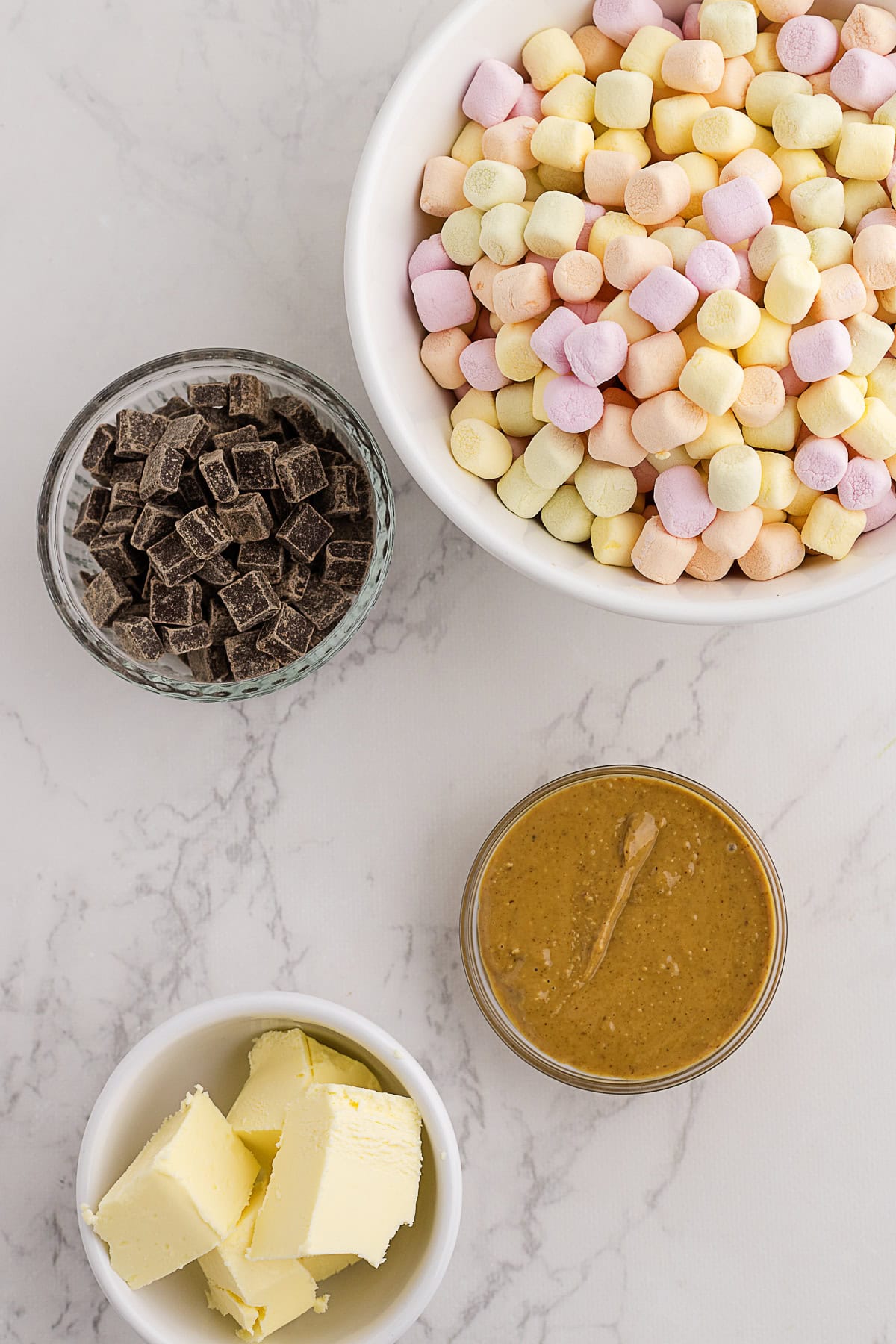 A bowl of mini marshmallows, a bowl of chocolate chunks, a bowl of butter, and a jar of peanut butter on a marble surface.