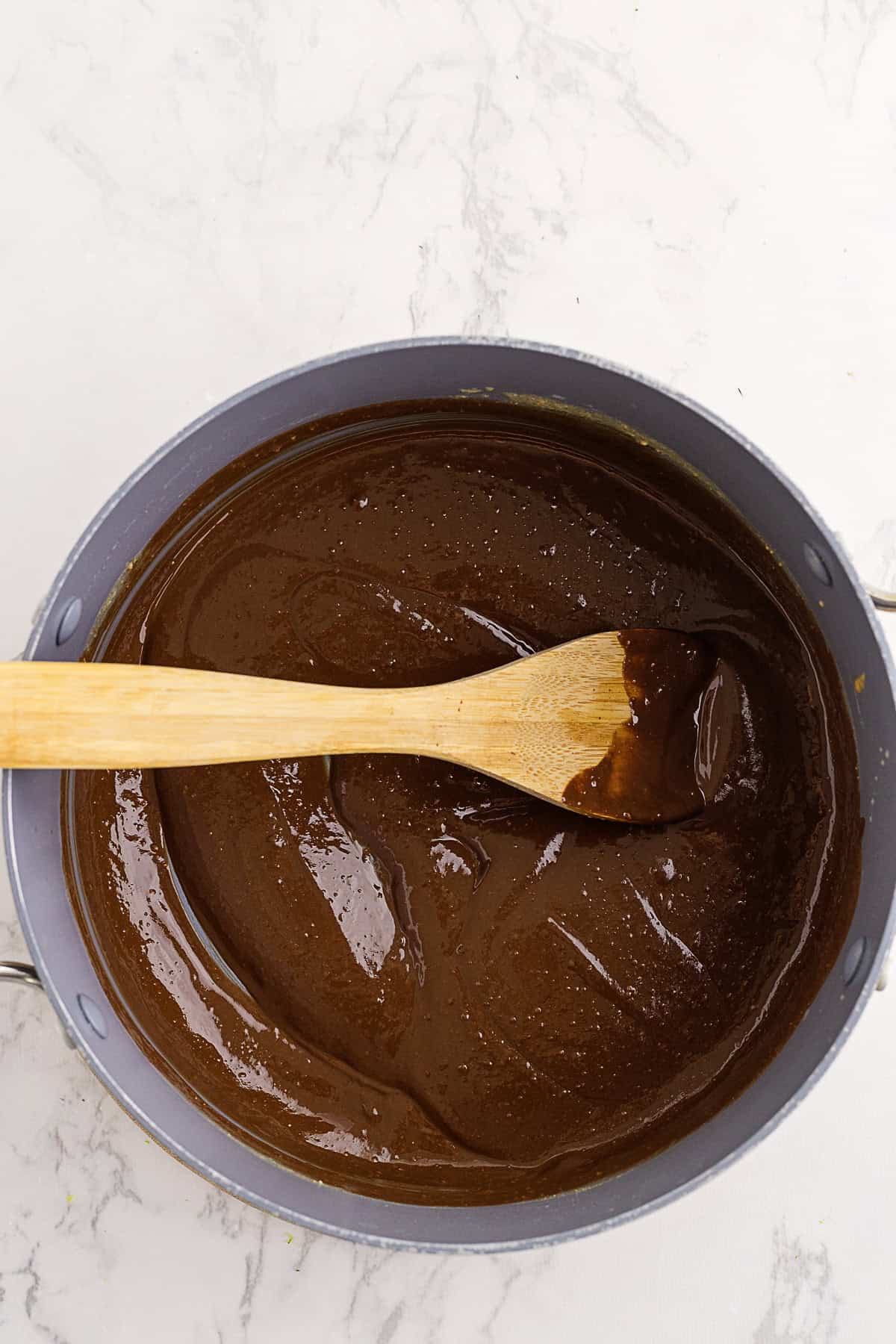 A wooden spoon rests in a pot filled with smooth, glossy chocolate batter on a light marble surface.