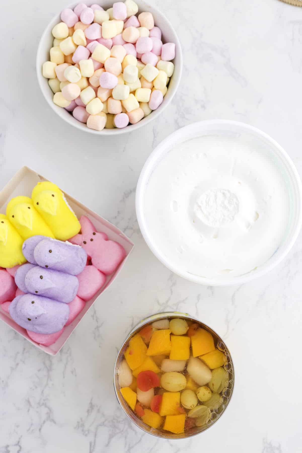 A bowl of mini marshmallows, a container of whipped topping, a box of marshmallow chicks, and a bowl of mixed fruit on a marble surface.