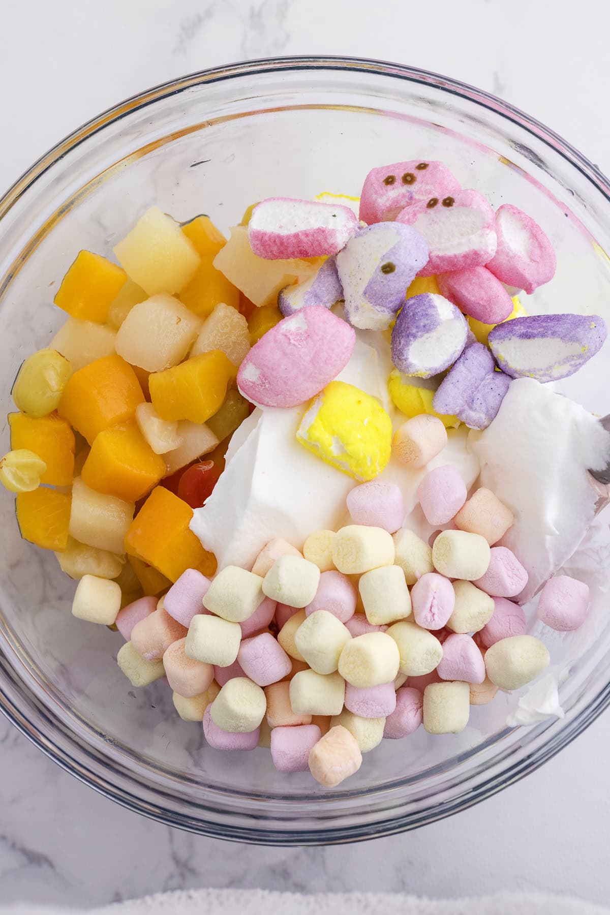 A glass bowl containing canned fruit, colorful mini marshmallows, marshmallow candies, and a large dollop of whipped topping.