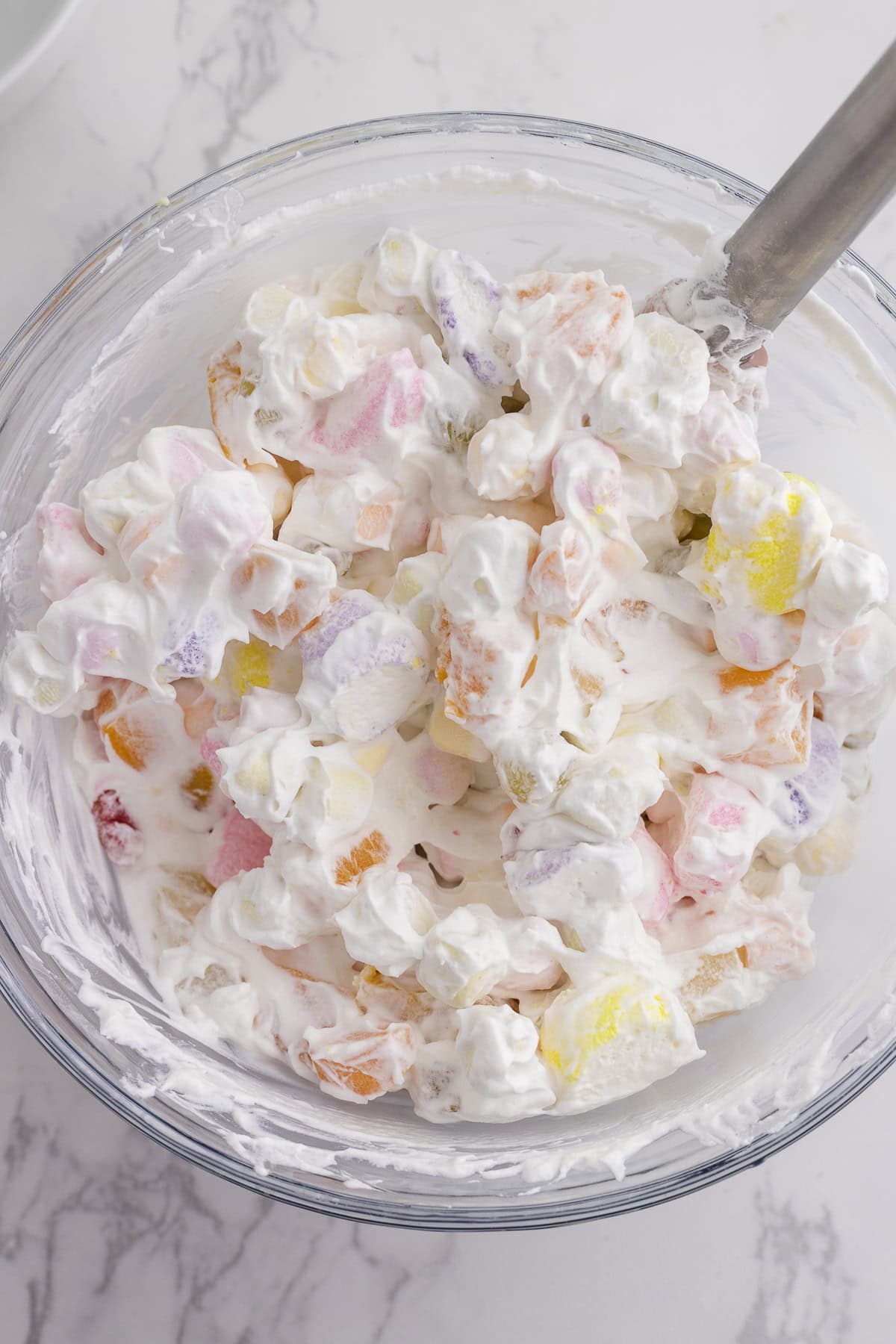 A glass bowl filled with a creamy fruit salad mixture containing colorful marshmallows and whipped topping, with a metal spoon inside.