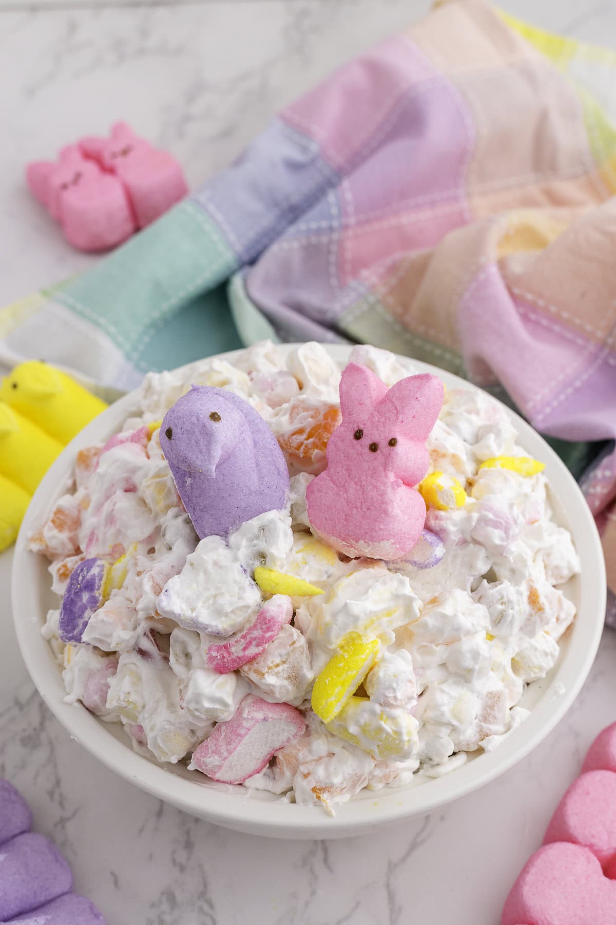 A bowl of marshmallow salad topped with pastel-colored marshmallow bunny and chick candies, with more candy and a multicolored cloth nearby.
